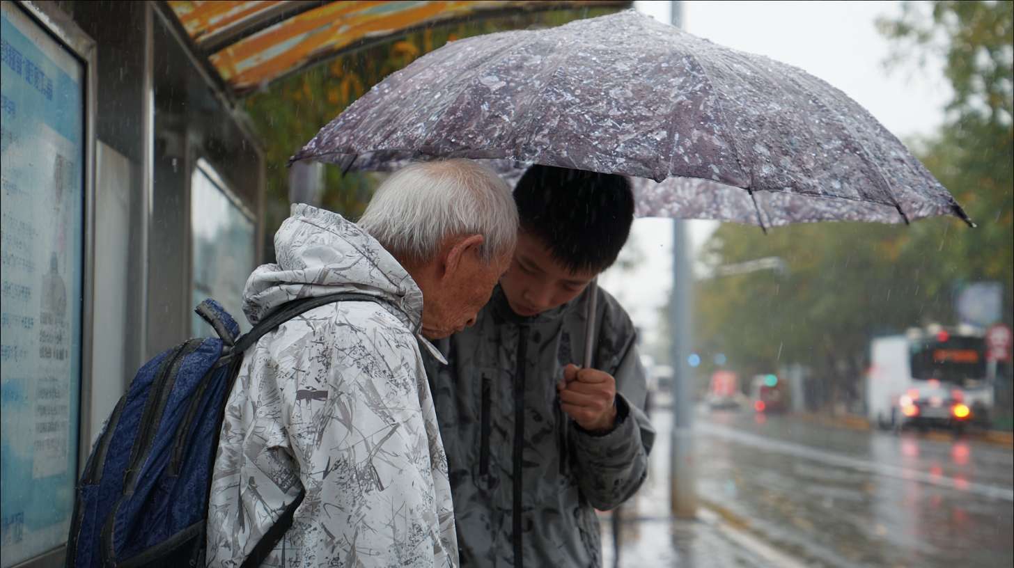 2. 白发老人、放学小男孩
• 地点：午后雨天的公交站台
• 事件：白发老人撑着小伞，伞沿倾斜护住身边背书包的小男孩，两人并肩站在雨棚下，老人不时调整伞的角度，小男孩偷偷把伞往老人那边推。 --ar 16:9 --v 7 --stylize 100 --fast --no 水印
