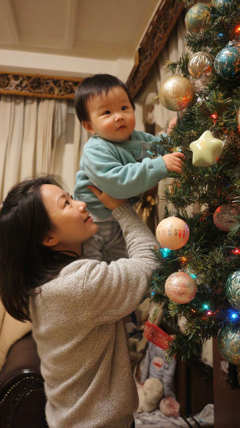 2圣诞树顶挂星星，妈妈托举孩子，树身缠绕彩灯，地面有未挂的袜子装饰，镜头从下往上推，温馨写实风 --ar 9:16 --v 7 --stylize 100 --fast --no 水印